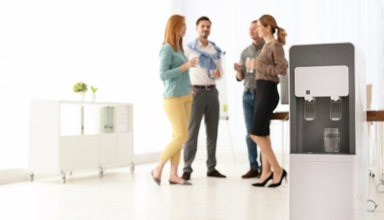 Modern water cooler with glass and blurred office employees on background. Space for text